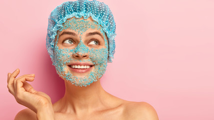 Headshot of pretty smiling woman looks with dreamy expression right, raises hand, focused aside, applies nourishing sea salt mask around face, wears waterproof showercap, has healthy soft skin