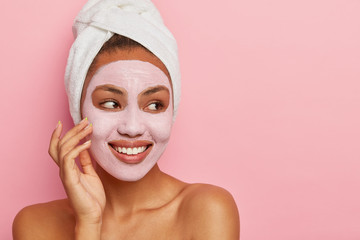Close up shot of lovely African American woman has delicate sooth skin, wears cream mask on face to reduce acnes, has healthy complexion, hygienic treatments wears white wrapped towel on head