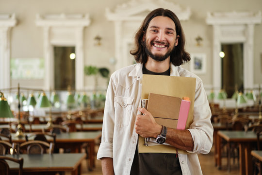Portrait of young attractive male student with book joyfully looking in camera in library of university