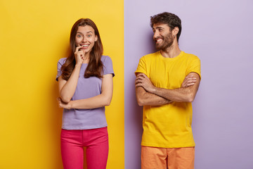 Fototapeta premium Indoor shot of positive young woman and man smile happily, being in good mood, spend free time together, wear t shirts, pose against purple background. People, relationship and emotions concept