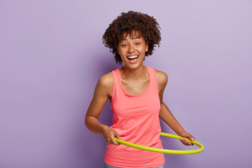 Happy girl with curly hairstyle, rotates hula hoop for slimming, has exercises in gym, smiles positively, wears pink vest, isolated over purple background. People, sport, lifestyle, motivation concept - obrazy, fototapety, plakaty