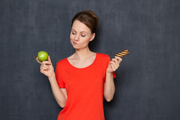 Portrait of doubting girl making choice between apple and cookie