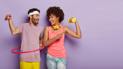 Horizontal shot of happy diverse couple have active sport training, raise dumbbells, rotate hula...