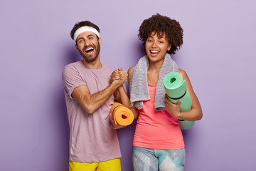 Indoor shot of happy diverse woman and man keep hands together, dressed in sportwear, hold fitness mats, meet together in gym before gymnastic exercises, laugh positively, isolated on purple wall