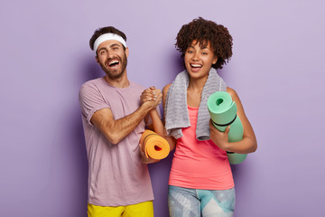 Indoor shot of happy diverse woman and man keep hands together, dressed in sportwear, hold fitness...