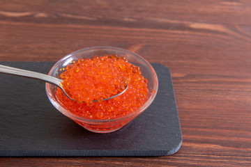 Red caviar in glass jar with silver spoon on the dark brown wooden background