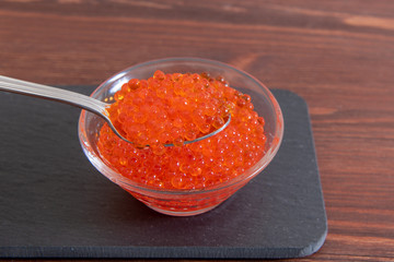 Red caviar in glass jar with silver spoon on the dark brown wooden background