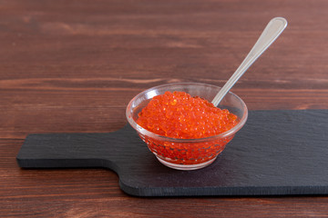 Red caviar in glass jar with silver spoon on the dark brown wooden background