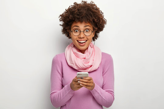 Studio shot of happy curly haired woman holds modern cellular, chats online, smiles joyfully, wears transparent glasses and casual violet jumper, poses indoor. Blogger addicted to social media