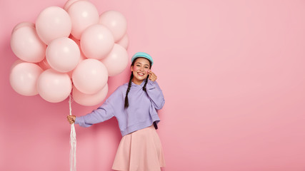 Positive lovely Korean woman smiles gently, shows like sign with hand, poses with airballoons, dressed in oversized sweater and skirt, celebrates special event, isolated over pink wall, copy space