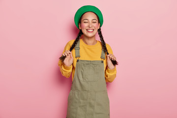 Cute modest teenage girl holds two pigtails, enjoys positive moment of life, wears green beret and sarafan, has piercing in nose, isolated over pink background, likes spending time in family circle