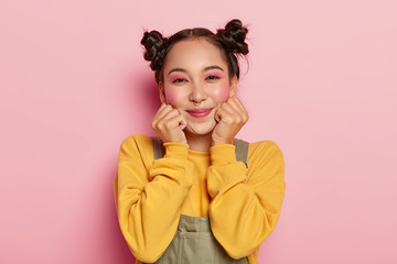 Portrait of happy Asian girl with pinup makeup, dark hair combed in two buns, piercing in nose, wears casual yellow sweatshirt and overalls, stands on pink background, feels happy. Ethnicity, emotions