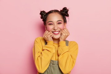 Obraz na płótnie Canvas Portrait of beautiful Asian girl with pinup makeup, holds chin with both hands, dressed in casual outfit, has dark hair combed in two buns, poses against rosy background, wears piercing in nose