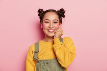 Headshot of cheerful lovely young Asian woman with rouge cheeks, keeps one hand under chin, has two buns, wears yellow sweater and brown overalls, poses against pink background. Emotions concept