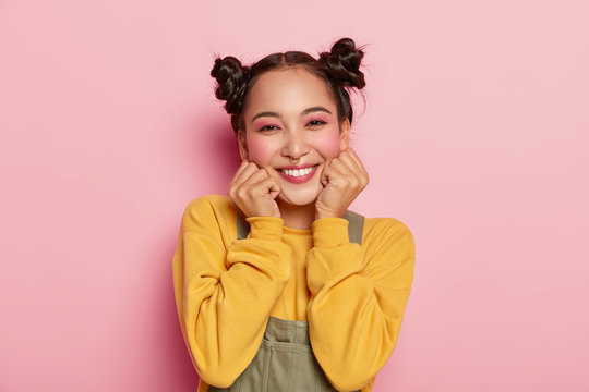Portrait of beautiful Asian girl with pinup makeup, holds chin with both hands, dressed in casual outfit, has dark hair combed in two buns, poses against rosy background, wears piercing in nose