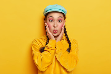 Amazed Asian lady keeps lips rounded, looks with surprise, reacts on gossip, wears stylish hat and corduroy yellow sweatshirt, has pink makeup, piercing in nose, poses indoor. Monochrome image
