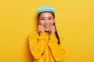 Pleasant looking cheerful Asian girl keeps both hands under chin, has pinup makeup, wears blue stylish cap, velvet yellow hoody, isolated, expresses positive emotions, has charming smile, two pigtails