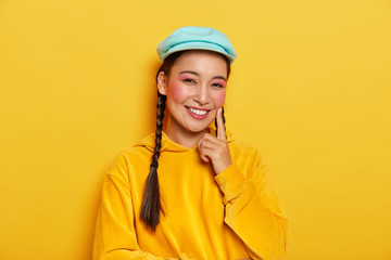 Positive brunette woman with healthy complexion, touches rouge cheek with index finger, smiles happily, dressed in casual yellow hoody, blue cap, enjoys free time for go shopping. Ethnicity.