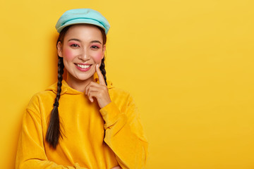 Emotions, ethnicity and happiness concept. Attractive Asian lady with pink makeup, keeps index finger on cheek, ponders on good idea, wears casual yellow velvet sweatshirt with hood, smiles gently