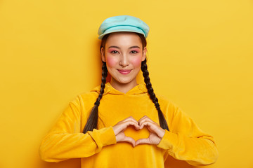 Be my valentine. Lovely young girl with asian appearance, shapes heart with both hands, has long hair combed in two plaids, rosy cheeks, wears piercing in nose, isolated over yellow background