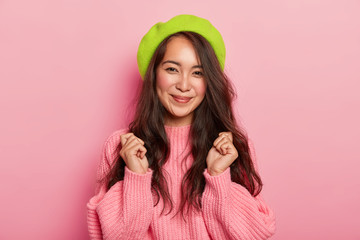 Joyous dark haired woman with pleasant smile, raises clenched fists, awaits something awesome, dressed in oversized sweater and beret, has natural beauty, isolated over pink studio background.