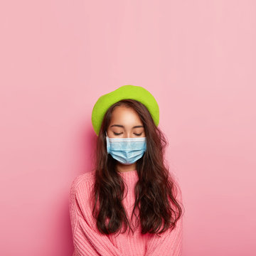 Vertical Image Of Calm Ill Woman Covers Nose And Mouth With Medical Mask, Has Infectious Disease, Wears Protective Mask In Public Place, Dressed In Fashionbale Headgear And Pink Knitted Sweater