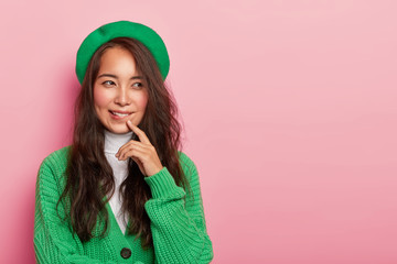 Thoughtful brunette woman wears green beret and jumper, bites lips, has contemplative look aside, tries to find decision, poses against pink studio wall with copy space. Face expressions concept - obrazy, fototapety, plakaty