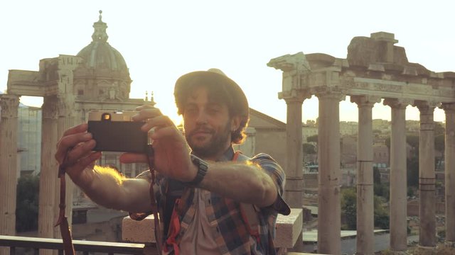 Young man tourist backpacker taking selfies with vintage camera at Roman Forum at sunrise. Historical imperial Foro Romano in Rome, Italy from panoramic point of view.