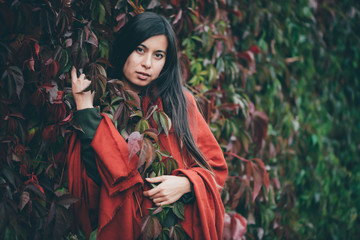 Dreamy beautiful girl with long natural black hair on autumn background of green red grape hedge. Inspired girl in red shawl and green coat. Autumn euphoria. Female emotional portrait in faded tones.