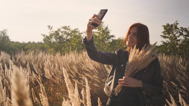 Attractive fun stylish redhead woman in the field at sunset making a selfie with a bouquet of flowers. Beautiful woman in a leather jacket