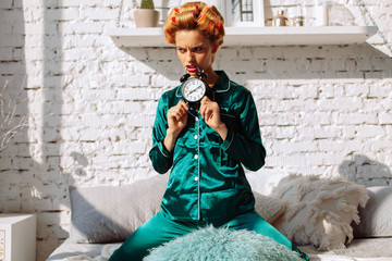 angry fashion woman in silk pajama bitting alarm clock in bedroom interior
