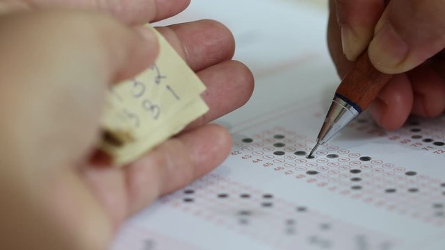 Education concept. Student's hands filling in standardized test form with black pencil drawing selected choice on answer sheets and cheating in test with answer piece of paper in hand. 