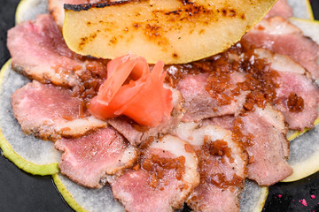 Edged pieces with caramelized pear on a black ceramic plate. Baked meat sliced with thin pieces