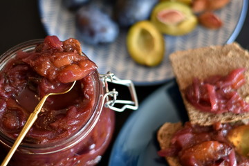 Plum chutney or jam in a glass jar with open lid and golden spoon on black tabletop. Slices of brown bread and plate with plums in blurred background. Selective focus. - obrazy, fototapety, plakaty
