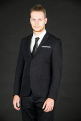 Portrait of young smart and handsome business man in black suit standing on isolated black background.