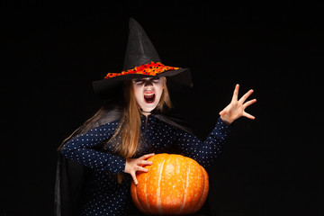 Halloween Witch with Pumpkin on black background Casts a spell with his hands. Beautiful young surprised woman in witches hat and costume holding pumpkin. Wide Halloween party art design. Copy-paste