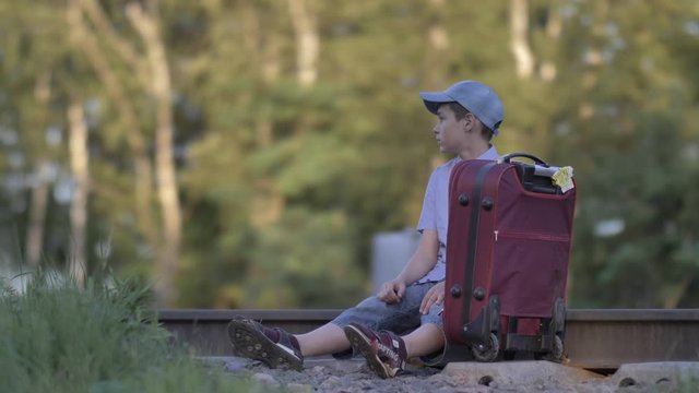sad boy in a cap sits on a suitcase on the railway outdoors