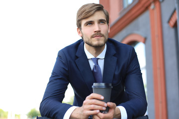 Pensive young businessman holding cup of coffee while standing with his bicycle near office building in the background.