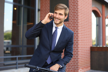 Portrait of a smiling young business man dressed in suit talking on mobile while standing with bicycle outdoors.