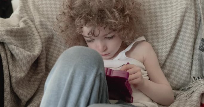 a cute little boy slouched back or reclining on a sofa with his knees up is drawing on a pad, he has long curly hair and wearing a singlet, tank top