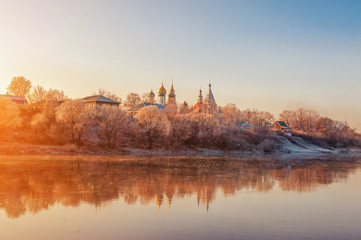 Winter landscape of temples of the city with sunset on the river Bank. The trees are covered with snow, the setting sun illuminates a beautiful mysterious landscape