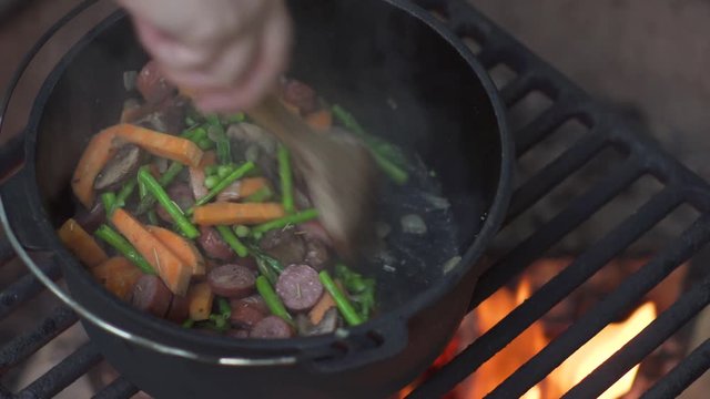 Close-up of woman lifting lid and stirring meal as it cooks over campfire in cast iron dutch oven