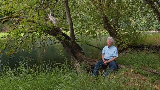 Older man sits on large tree root near river and looks into camera