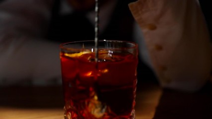 Bartender's hands mixing an alcoholic drink with a large piece of ice in glass using a mixing spoon on a wooden bar table in light motion captured in 120fps slow motion.