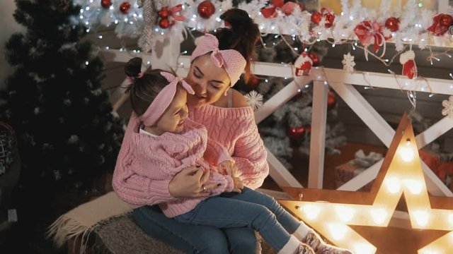 Happy mother having fun with her kid daughter near Christmas tree. Beautiful mother and daughter in pink pullovers. Mother tickling her little daughter in slow motion. Motherhood concept. Medium shot