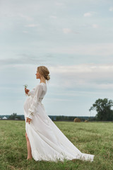 Pregnant girl walks in a field near haystacks in a long white dress, a woman smiles and holds her...