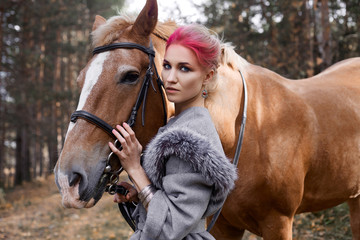 Fototapeta premium Woman on a horse in the fall. Creative bright pink makeup on the girl face, hair coloring. Portrait of a girl with a horse. Horseback riding in the autumn forest. Autumn clothes