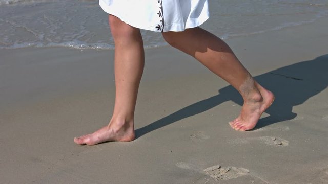 Woman legs in white dress walking barefoot alone on the beach