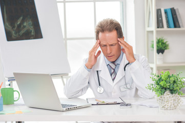 Senior doctor at his office in hospital working using laptop headache