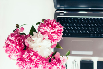 bouquet of pink peonies, laptop, smartphone, pens, glasses and a notebook on a white table, top view.